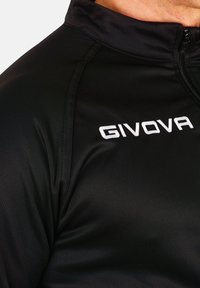 Črna športna jakna iz gladkega, lahkega materiala; ima stoječ ovratnik in bel "GIVOVA" logotip na levem prsnem delu.