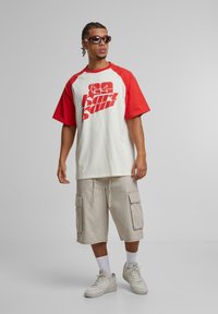 Red and cream oversized t-shirt with graphic print, beige cargo shorts, white sneakers, and sunglasses, displaying a casual, urban style.