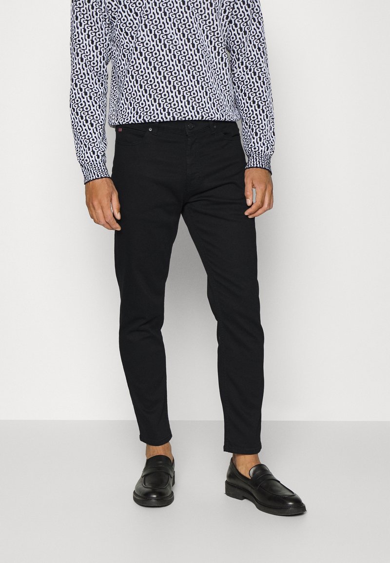 Uomo che indossa pantaloni neri slim-fit, mocassini neri in pelle e una camicia a maniche lunghe con un motivo astratto bianco e nero.