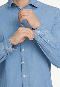 Vaaleansininen farkkupaita, jossa on nappihaalarit. Varustettu klassisella kauluksella ja etupaneelilla, jossa on näkyviä nappeja ja sileä pinta.