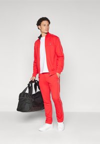Chándal rojo con acentos blancos, chaqueta con cremallera y pantalones ajustados. El modelo sostiene un bolso de lona negro y lleva zapatillas blancas. Textura suave.