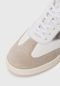 Sneaker en cuir blanc avec des accents en suède beige et un détail en argent brillant. Équipé de lacets plats blancs et d'un design perforé à l'avant.