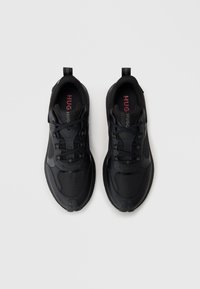 Black athletic shoes with textured panels, lace-up design, and a pull tab. Features bold "HUG" branding on the insole.