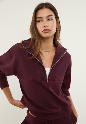 Felpa bordeaux con zip e collo a camicia, polsini a coste e vestibilità comoda. Abbinata a un top bianco sotto. Texture liscia.