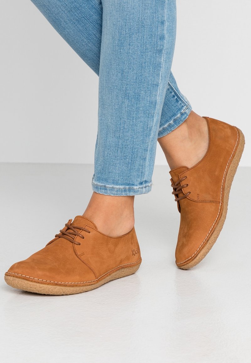 Des pieds dans un jean bleu clair portant des chaussures décontractées en cuir marron à lacets sur un sol blanc, avec une jambe légèrement relevée.