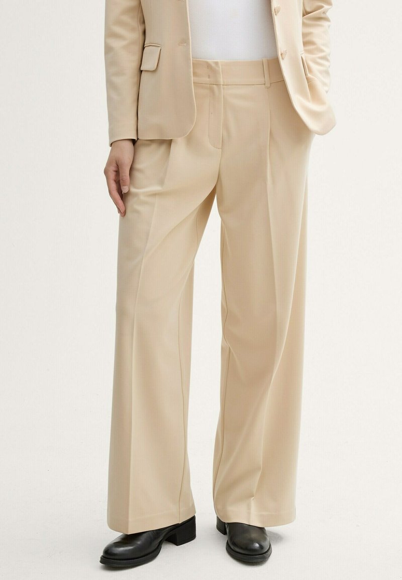 Pantalones de pernera ancha color beige, confeccionados en una tela suave con un ajuste a medida. Presentan bolsillos delanteros y una cintura de altura media. Combinados con zapatos negros.
