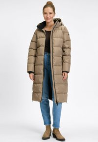 Frau in einem beigen langen Puffermantel mit Kapuze, blauen Jeans, schwarzem Oberteil und beige Wildleder-Stiefeletten, stehend vor einem weißen Hintergrund.