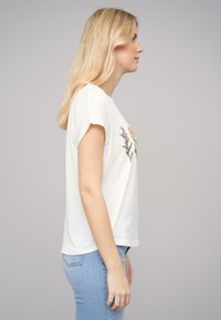T-shirt à manches courtes blanches avec broderie florale dans des couleurs neutres. Le modèle est associé à un jean bleu clair, mettant en avant une coupe décontractée.