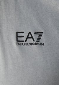 Logo noir EA7 Emporio Armani brodé sur tissu gris clair.