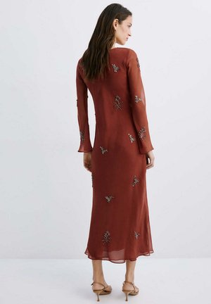 Mujer con cabello largo y oscuro que lleva un vestido de manga larga color óxido adornado con pequeños patrones de cuentas florales, de pie frente a un fondo liso.