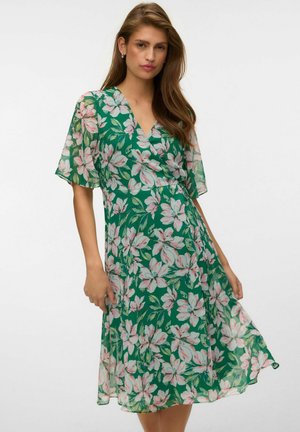 Femme aux longs cheveux bruns portant une robe portefeuille verte à motifs floraux avec des manches courtes à volants sur fond blanc.