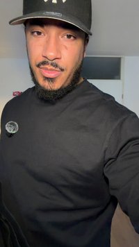 Sudadera negra con cuello redondo y tejido texturizado, con un pequeño micrófono gris de clip en el pecho izquierdo. Se lleva con una gorra de béisbol negra.