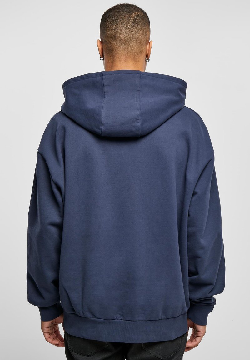 Personne aux cheveux courts portant un sweat à capuche bleu marine ample, vue de dos sur un fond gris clair uni.