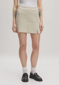 Beige geweven minirok met een lichte voorknip, twee zijzakken en een gestructureerde afwerking, gecombineerd met zwarte instappers en witte sokken.