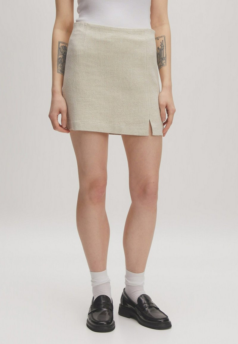 Beige geweven minirok met een lichte voorknip, twee zijzakken en een gestructureerde afwerking, gecombineerd met zwarte instappers en witte sokken.