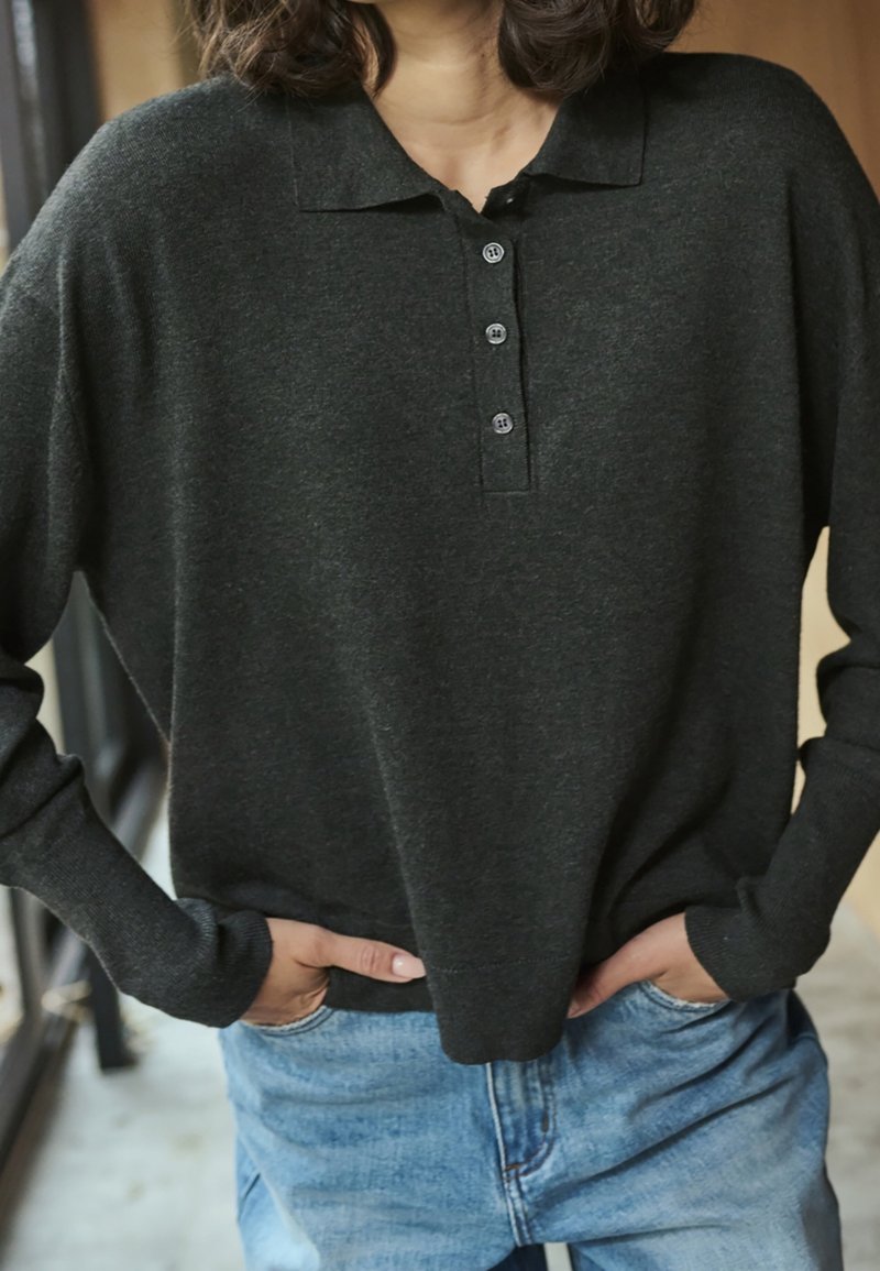 Personne portant un polo à manches longues gris foncé et un jean bleu, les mains dans les poches, debout à l'intérieur près d'une fenêtre.