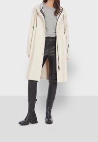 Imperméable beige clair avec capuche, boutons-pression et cordons, associé à un pantalon en similicuir noir et des bottes noires montantes jusqu'aux genoux.