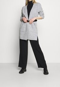 Szary dziergany kardigan z głębokim dekoltem w serek, otwartym przednim panelem, dwoma kieszeniami i paskiem w talii, zestawiony z czarnymi spodniami o szerokich nogawkach i botkami.