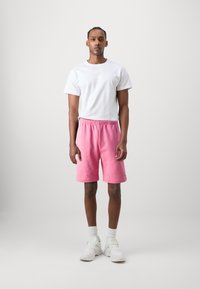 Różowe bawełniane szorty z elastycznym pasem i bocznymi kieszeniami, zestawione z białą koszulką z okrągłym dekoltem i białymi butami sportowymi.
