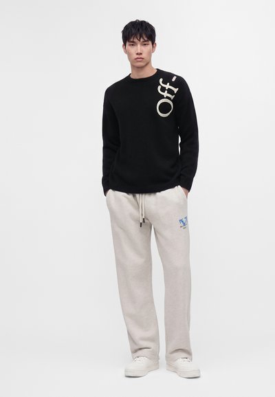 Jeune homme debout portant un pull noir avec le texte "Off" et un pantalon de survêtement gris clair avec un logo bleu, ainsi que des baskets blanches sur un fond uni.