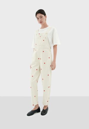 Salopette blanche avec broderie florale rouge, coupe décontractée, accents en boutons et poches latérales, associée à un t-shirt blanc et des chaussures noires.