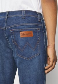 Tumed sinised denim teksad, millel on hele pruuni nahkplekk, kus on kirjas "Wrangler." Tunnuslikud tagataskute õmblused W-kujulised.