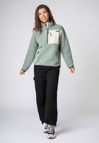 Fleece pullover in lichtgroen met een crèmekleurige voorzak, ritsluiting en twee ritszakken aan de zijkant; gecombineerd met zwarte cargo-broeken.