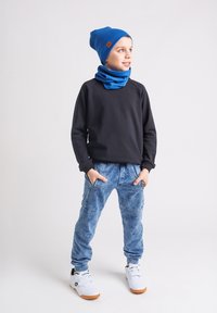 Dziecko w niebieskiej czapce i kominie, czarnej bluzie z długim rękawem, niebieskich spodniach dresowych i białych sneakersach, z rękoma w kieszeniach, patrzące w prawo.