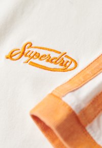 Hvid bomulds T-shirt med et broderet orange "Superdry" logo og orange stribede detaljer på ærmerne. Blød tekstur, afslappet design.