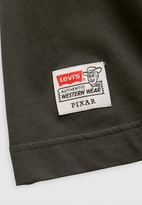 Tejido gris oscuro con un parche de logo de Levi's que presenta "AUTÉNTICA ROPA DEL OESTE" y una ilustración de un personaje de Pixar. Detalle de costura visible.