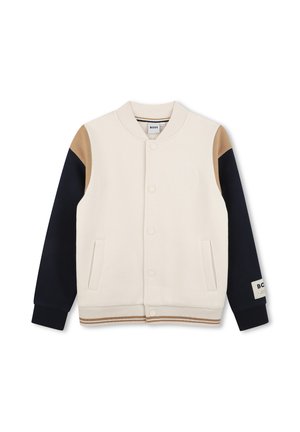 Beige varsity-stijl jas met marineblauwe mouwen en bruine schouderstukken, vooraan drukknoopsluiting en twee voorzakken.