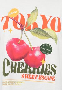 Λευκό μπλουζάκι με γραφική εκτύπωση δύο κόκκινων κερασιών με πράσινα φύλλα, τολμηρό πορτοκαλί "TOKYO" και κυκλικό ετικέτα με την επιγραφή "Juicy Red Sakuranbo Delight".