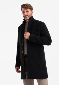 Abrigo negro de lana con un alto cuello, cuenta con seis botones, bolsillos laterales y un corte ajustado, usado sobre un suéter claro y pantalones beige.