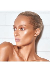 Profil av en modell med jevn, bronsert hud, fremhevede kinnbein, lys rosa lepper og lyse øyne mot en hvit murvegg.