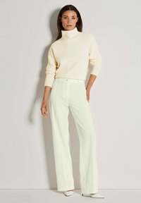 Vrouw die tegen een witte muur staat in een crèmekleurige geribbelde coltrui, witte high-waisted wijde broek en witte hakken.