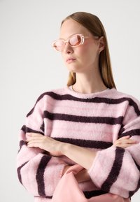 Svetr vyrobený z načechraného růžového materiálu s tmavě fialovými pruhy. Módní oversize střih se širokými rukávy. Modelka nosí sluneční brýle s růžovým nádechem a růžový doplněk.