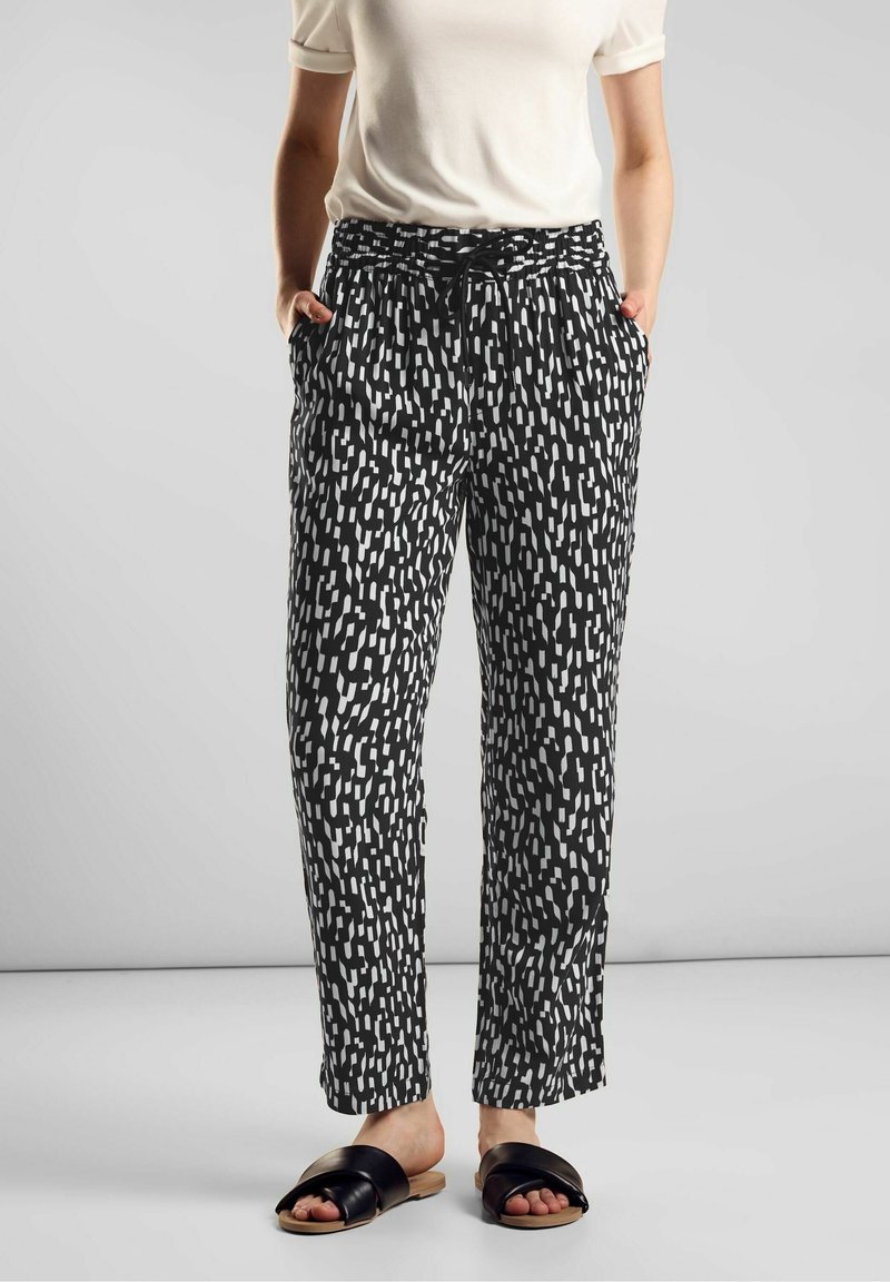 Personne portant un pantalon à motifs noir et blanc, un haut blanc à manches courtes et des sandales noires ouvertes à enfiler, se tenant avec les mains dans les poches.
