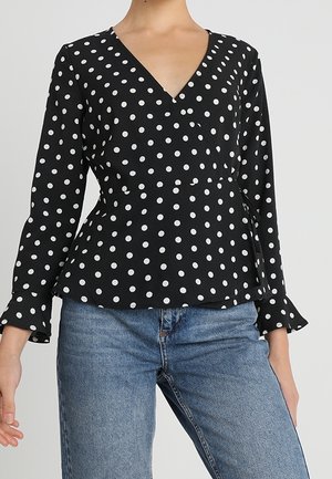 Kvinde iført en sort, langærmet slå-om bluse med hvide polkaprikker og blå højtaljede jeans, stående foran en ensfarvet baggrund.