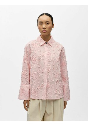 Ženska, ki nosi svetlo roza srajco z gumbi in cvetlično vezenino ter bež ohlapne hlače, stoji na enobarvnem belem ozadju.