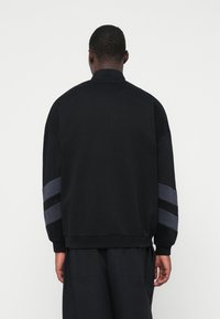 Felpa nera con collo alto e spalle scese. Presenta strisce orizzontali grigie e nere sulle maniche. Tessuto morbido.