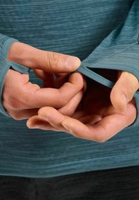 Langsleeve, teal sportshirt met duimgaten. Close-up van handen die de manchetten aanpassen, waarbij de soepele textuur en het geïntegreerde ontwerp worden getoond.