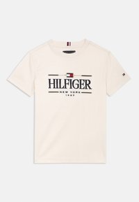 T-shirt en coton crème avec un grand logo "HILFIGER" en marine, rehaussé d'accents rouges et bleus, col rond et manches courtes. Design minimaliste.
