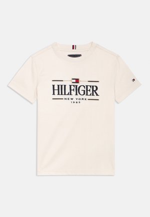T-shirt in cotone crema con grande logo "HILFIGER" in blu navy, con dettagli rossi e blu, colletto a girocollo e maniche corte. Design minimalista.