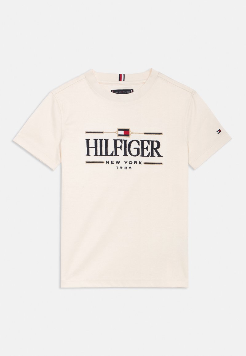 T-shirt en coton crème avec un grand logo "HILFIGER" en marine, rehaussé d'accents rouges et bleus, col rond et manches courtes. Design minimaliste.