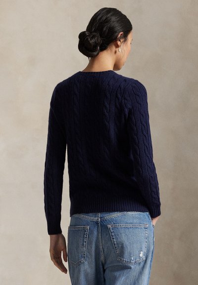 Femeie cu coafură coc închis la culoare poartă un pulover albastru închis cu model împletit și blugi albastru deschis, stând în fața unui fundal bej simplu, cu fața întoarsă.