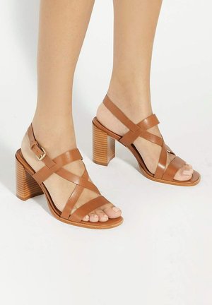 Brown leather sandals with cross-strap design, open heel, and a textured block heel. Buckle closure on ankle strap.