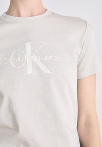 Svetlošedé bavlnené tričko s krátkymi rukávmi, s veľkým bielym logom "CK" a pod ním v subtílnom, embosovanom štýle textom "Calvin Klein Jeans".