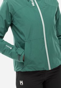Veste imperméable verte avec une fermeture éclair à l'avant, des poignets réglables et des accents de logo. Présente une texture douce et un design ajusté.