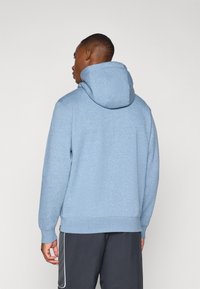 Lys blå hoodie med et struktureret, blødt stof. Har en stor hætte og ribbede manchetter, båret med mørkegrå bukser. Bagansigtet vises.