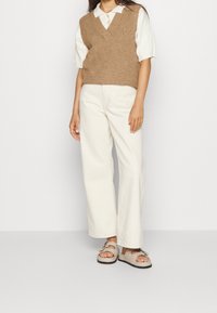 Bruin gebreid vest met een V-hals over een wit shirt met korte mouwen, gecombineerd met crème wijd uitlopende broek en beige sandalen met decoratieve accenten.